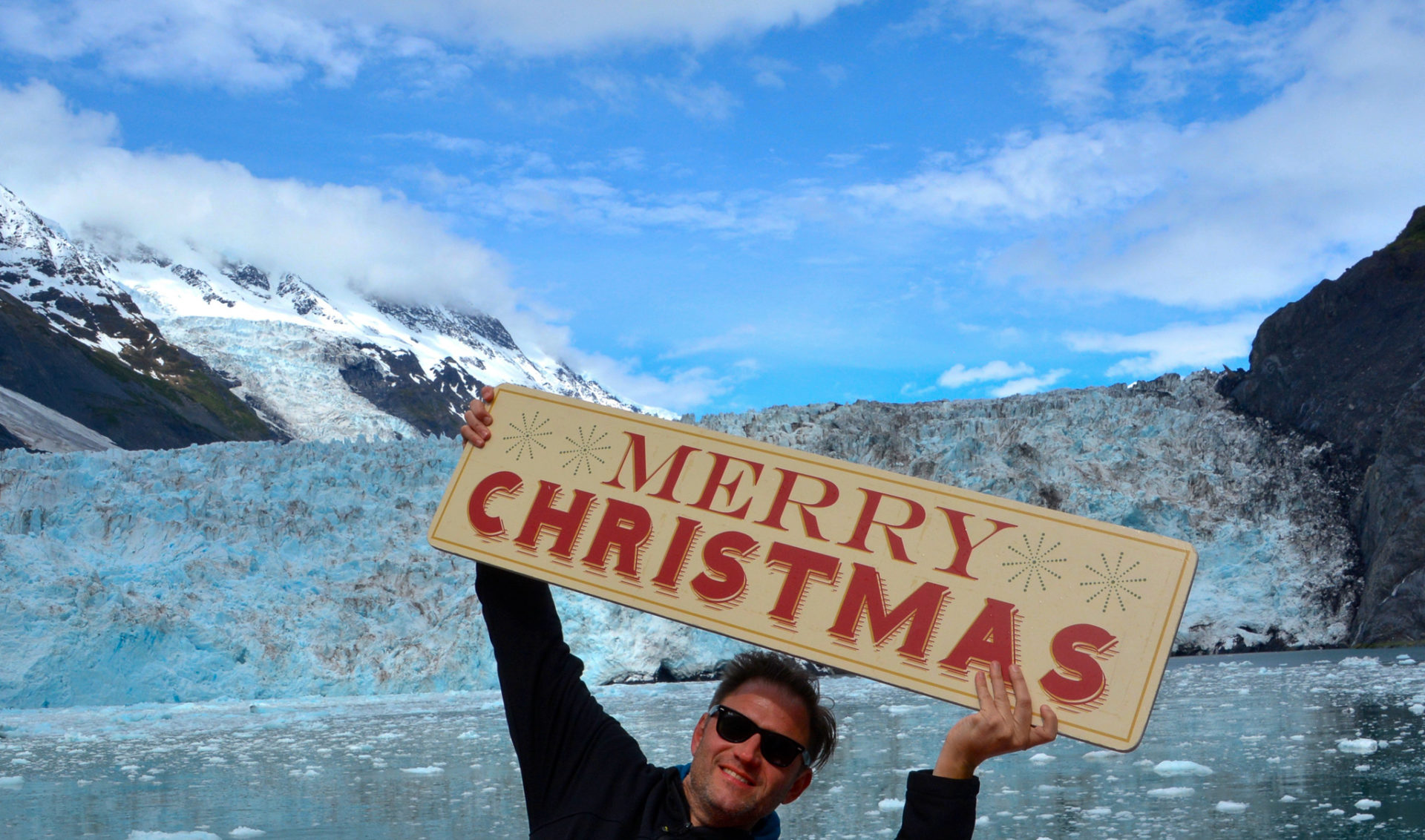 Autor Ralf Johnen hält vor einem Gletscher in Alaska ein Schild mit der Aufschrift "Merry Christmas" hoch