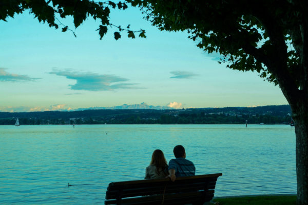 Pärchen sitzt auf einer Bank am Bodensee und schaut auf die leuchtenden Alpen im Hintergrund