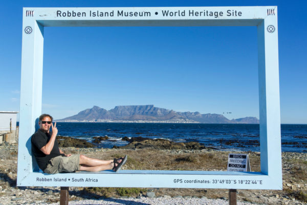 Autor Ralf Johnen sitzt auf Robben Island in einem Bilderrahmen, der den Tafelberg und das Meer einrahmt