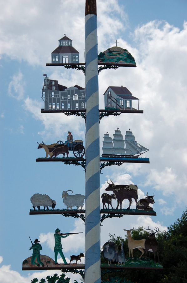 blauweiß geringelter Mast mit Motiven wie Segelschiff, Schafen, Kirchen, Hügeln und Wild