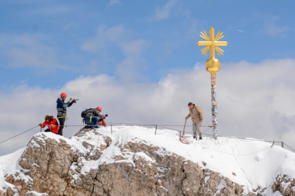 Vier Männer mit Kameras und Alpinausrüstung mit Gipfelkreuz auf der Zugspitze