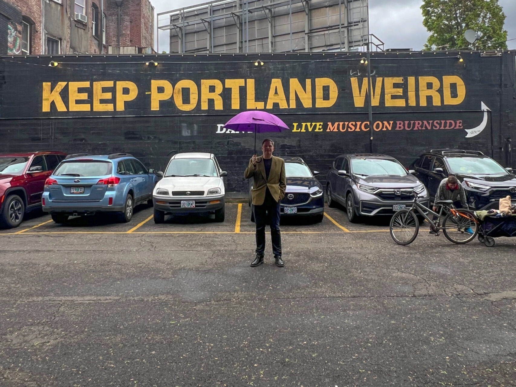 Autor mit Regenschirm vor einem Mural mit der Aufschrift "Keep Portland Weird"